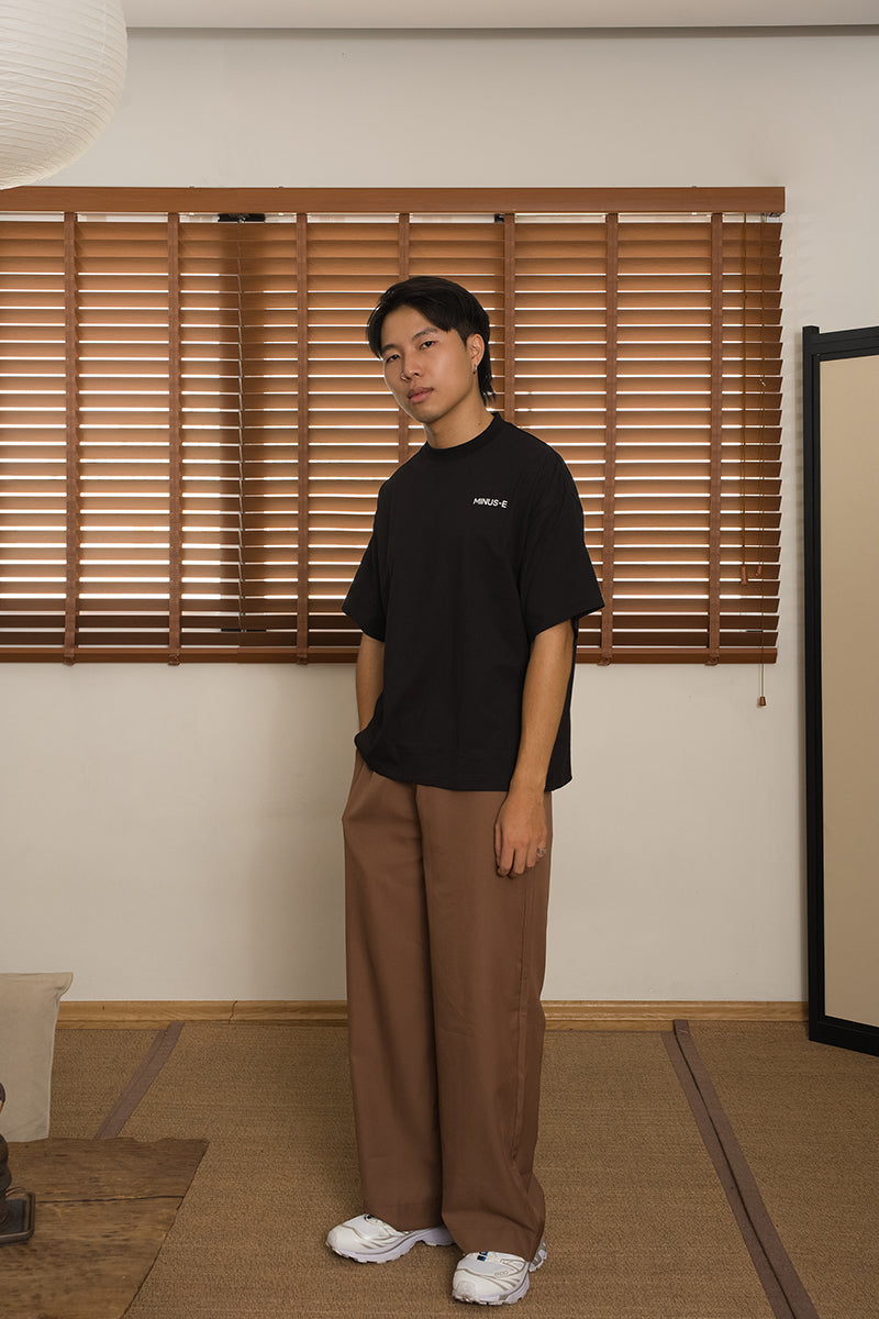 Person wearing a black t-shirt and brown pants from the brand MINUS-E standing in a room with wooden blinds. 