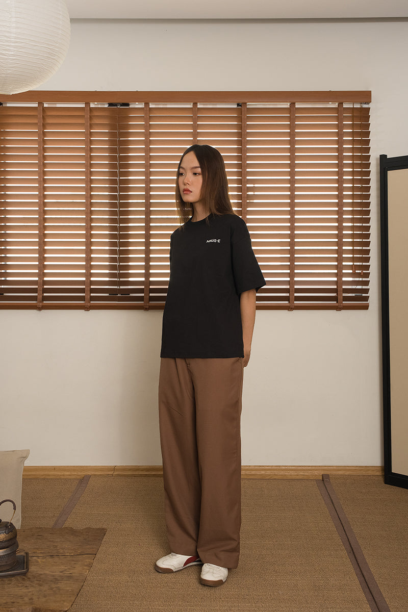 Woman wearing a black t-shirt and brown pants from MINUS-E standing in a room with wooden blinds.
