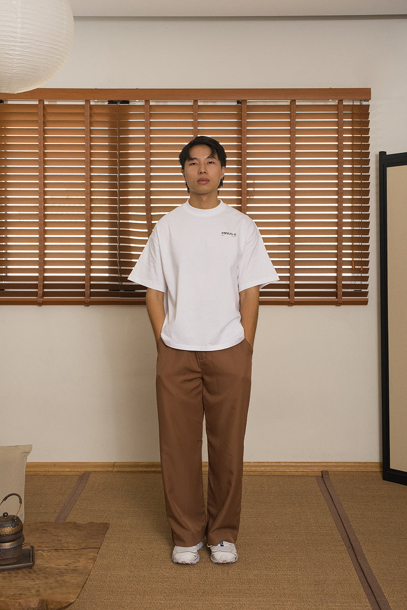 Male wearing a white t-shirt and brown pants, from the brand MINUS-E, standing in a room with wooden blinds and a traditional Japanese divider and floor.