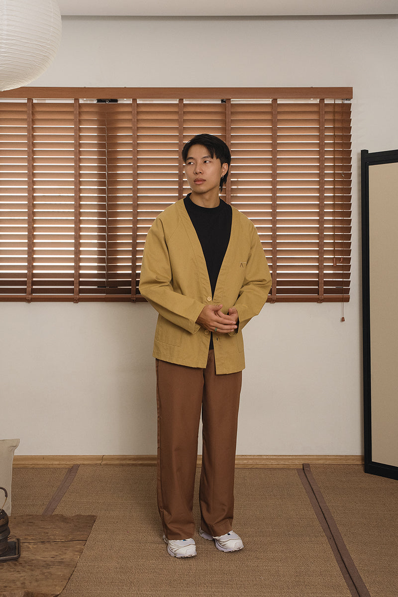 Men wearing a khaki kimono and brown pants standing in a room with wooden blinds. By MINUS-E.