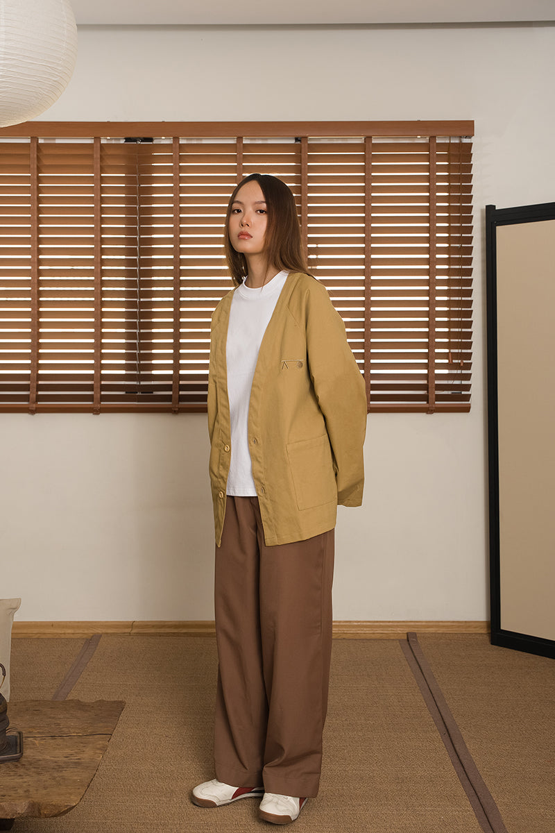 Person wearing a khaki kimono-inspired cardigan, designed by MINUS-E, and brown pants standing in a room with wooden blinds and a tatami mat floor.
