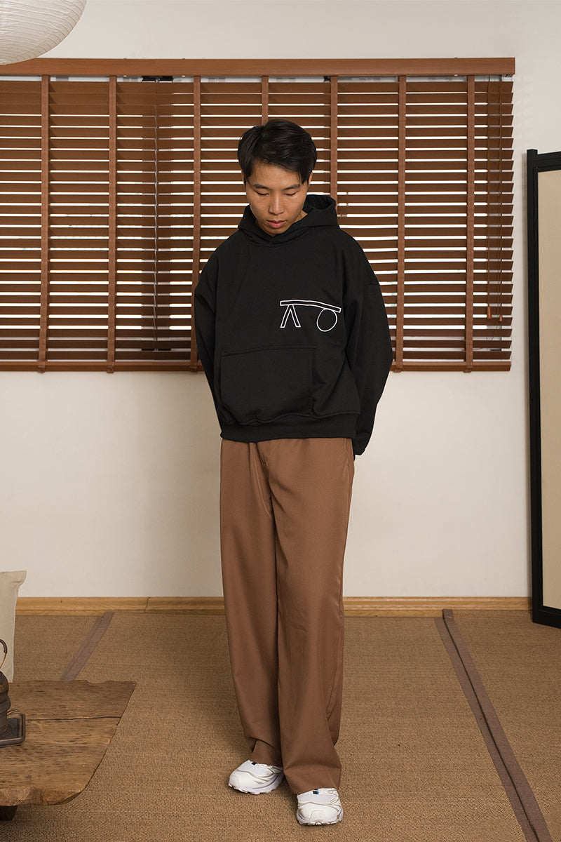 A male wearing a black hoodie with a white logo and brown pants standing in a room with wooden blinds. Designed by MINUS-E.