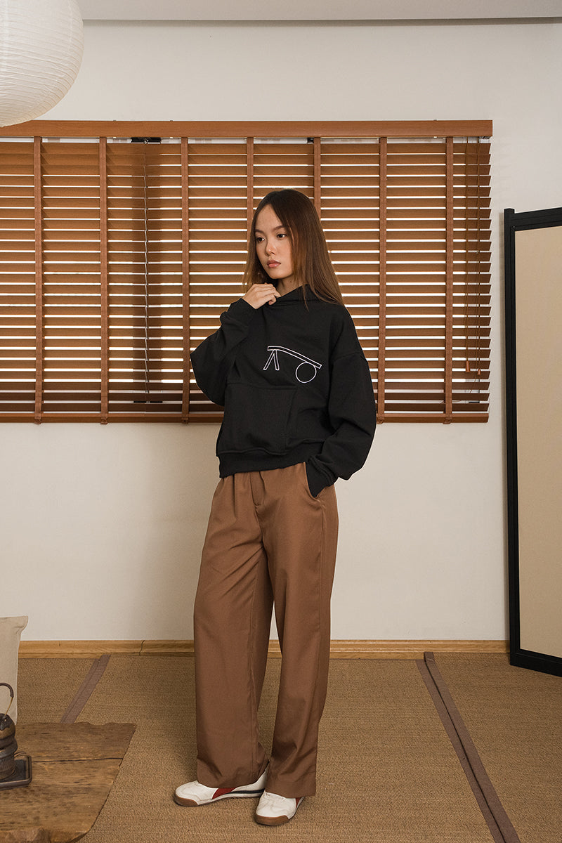 Woman wearing a black hoodie and brown pants standing in a room with wooden blinds and a white wall. Hoodie by MINUS-E.
